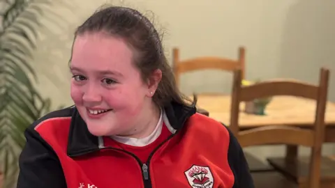 A girl sits in a cafe. She is wearing a school PE uniform in black and red, with a school crest on the front. Her long dark hair is in a ponytail and she is smiling widely. Here eyes look glassy and her face is flushed. Behind her there are some chairs and a table and a green long leafed plant. 