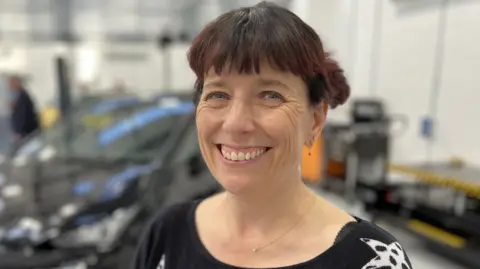 Alex Stroud standing in the centre smiling with thick brown hair tied up. Behind her, but not in focus, are electric vehicles parked up