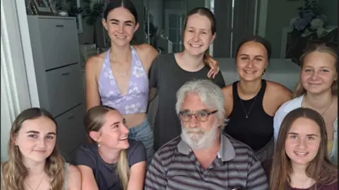 Foto de família Homem idoso, com cabelos e barba grisalhos, vestindo camiseta listrada preta e cinza, short preto e óculos. Ele está cercado por sete jovens vestidas com roupas de verão. Todo mundo está sorrindo e olhando para a câmera.
