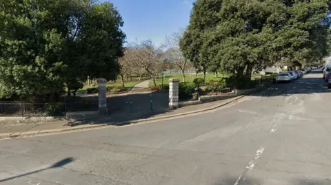 Google Entrance to Devonport Park. Two pillars either side of the park where people can walk through. There is a junction on the road outside with two big trees either side of the walkway. 