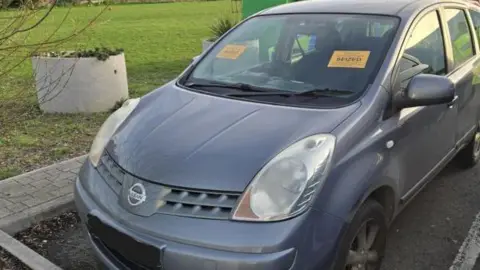 A grey Nissan Note car, which is parked in a car parking bay. Next to it is green grass and planters. On the car's window are two yellow stickers that read "seized" in black writing.
