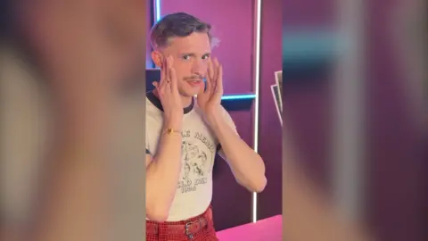 A man wearing a white t-shirt and red trousers stands with his hands against his face.