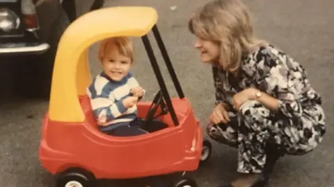 Family handout David as a young child. He is smiling at the camera and is sat in a children's red plastic toy car with a yellow roof. He is wearing a blue and white striped jumper and dark blue trousers. His mother is kneeling alongside the toy car, wearing a black and white floral patterned jumpsuit.