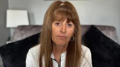 BBC/Emma Glasbey Lorraine Fraser has long, straight hair pulled back at the top with a hair tie, and the rest of the hair falls down past the shoulders. She is wearing a light-colored top with a zip at the front and a small microphone clipped near the neckline. A hoop earring is visible on one ear.
She is sitting on a large, dark upholstered chair with a soft texture.