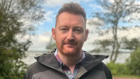 Dr Neil Reid looks into the camera. He has an auburn beard and short brown hair and is wearing a blue-purple shirt and a grey raincoat. A lake is behind him.