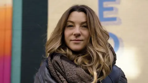 A woman with long brown hair stands in front of a colourful building. She is wearing a dark jacket and a chunky brown scarf wrapped around her neck.