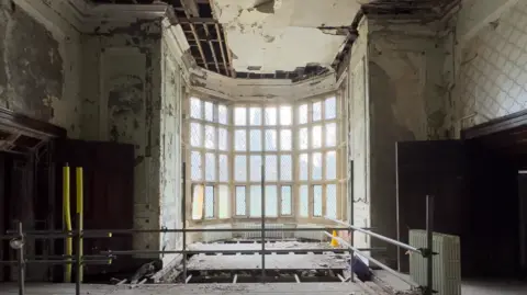 Hopwood Productions Inside one of the derelict rooms in Hopwood Hall. The image shows a large bay window, scaffolding, and walls stripped of paint and wallpaper. 