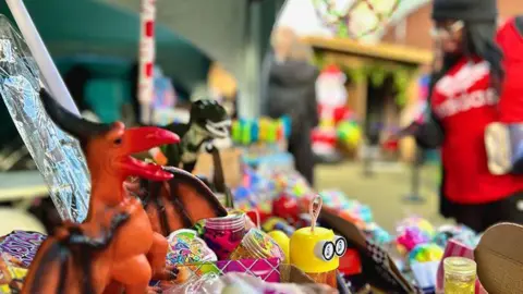 Erdington BID A plastic toy bird, a minion toy and other small toys. In the background is someone near the stall and further away a shape of a Father Christmas is in the background. 