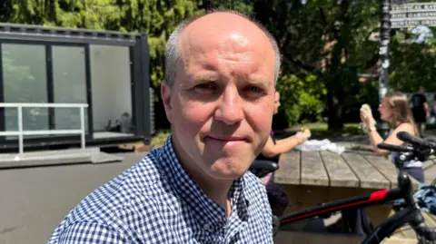 Arek is an older, balding man with short, grey hair. He is smiling at the camera with his mouth closed. He is wearing a blue and white checkered shirt. In the background, there are two people sitting at a picnic bench behind him.