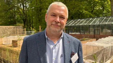 Ed Hanson/BBC James Cox looking into the camera with a slight smile. He has short grey hair and a grey beard and is wearing a blue blazer with a lighter blue shirt underneath. He is standing in front of a garden with raised beds covered in clear material. There is a greenhouse behind the garden with trees surrounding it.