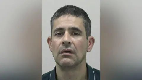 Mugshot of Andreas Michael. He has a wonky mouth, faint stubble and short dark hair tinged with grey.