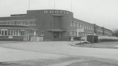 A general view of the Hoover factory. The brown building has 'Major Appliance Division' on two sides of it in white lettering. The entrance building is rounded with steps leading up to it. Two cars can be seen in the courtyard in front of it, as two people walk past on the street in front of it. 