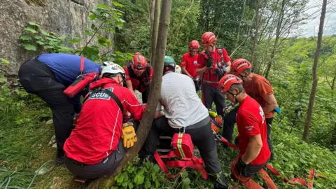 Edale MRT Rescuers surrounding a casualty at the foot of a rockface, in a wooded area