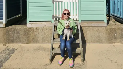 Becky Excell Becky holding her dog on her lap, sitting down in front of a beach hut