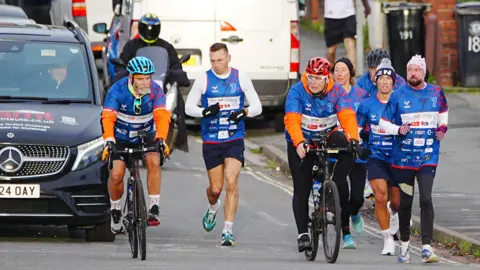 PA Media Kevin Sinfield and his support team running through residential streets. He is flanked by three people on bicycles and three other runners, while a support car trails behind.