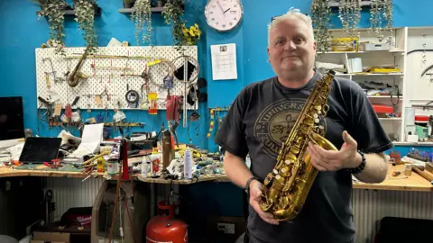 Amy Monkman/BBC A man in a black t-shirt with white hair holding a saxophone in what looks like a workshop. It has blue painted walls and tools and a laptop on the desk. 