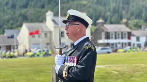 Cunningham, who is wearing the black and white IOM Coastguard uniform and peaked cap, carrying the sword, which has a wooden handle.