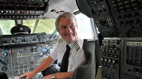 A pilot, Mike Bannister, sat in a plane cockpit and looking behind him. He is looking beyond the camera.