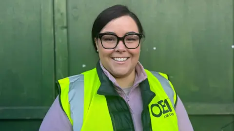 Emma Goodman - a woman with dark hair tied back, wearing black square glasses, a purple zip-up hoodie and a yellow hi-vis jacket smiles at the camera while standing in front of green wooden doors.