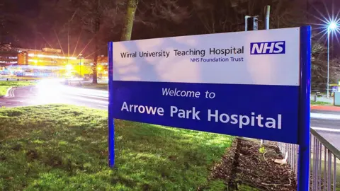 PA Media A sign in blue and white saying 'Welcome to Arrowe Park Hospital' it is evening and the hospital is lit up in the distance to the left