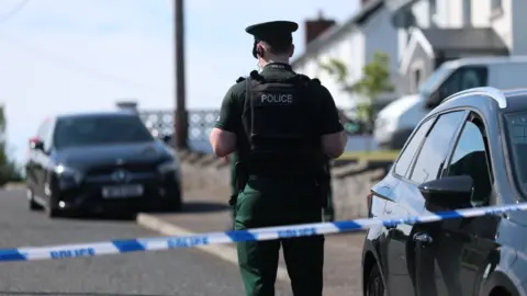 PA Media A police officer in full uniform is standing behind a blue and white cordon. This image is in a residential area. There are houses on the right and cars parked outside some o f them. 