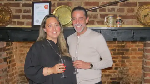 Sam Davies has long brown hair and is wearing a black blouse, standing next to David Wheeler, who is also holding a flute glass. They are both smiling inside, with a bricked fireplace behind them.