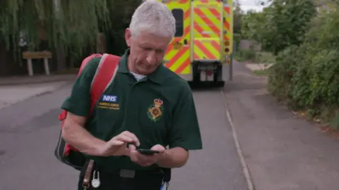 An NHS paramedic walking along a street, away from an ambulance, reading his mobile phone.