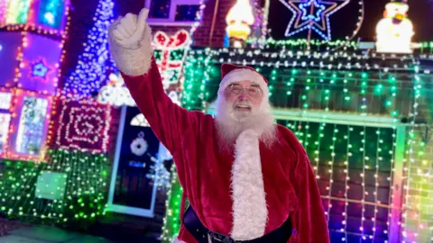 Anthony O'Toole dressed as Santa in front of his brightly-lit house which is covered in lights