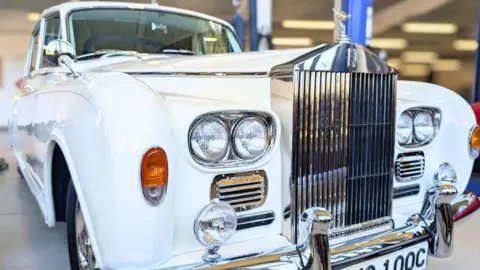The front of a white Rolls Royce in an indoor space with a large silver grill and the Rolls Royce symbol on top of it.