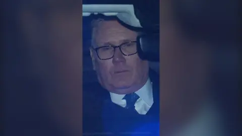 Sir Keir Starmer is seen in the back of a car. He looks weary. Blue lights from his motorcade flash against his face.