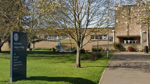 A google maps image of the front of Bottisham Village College. There is a pathway leading to the front doors of the building which is made from a light brown coloured brick. Next to the pathway is a grassy area with a sign for the collage and trees behind it. There is also a blue bike shed at the end of the grassy area, just in front of the school building. 