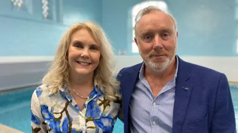 Yvonne Farmer, wearing an ocean blue and white shirt, has blond hair and smiles at the camera. On the right of her is her husband Trevor, wearing a blue suit. 