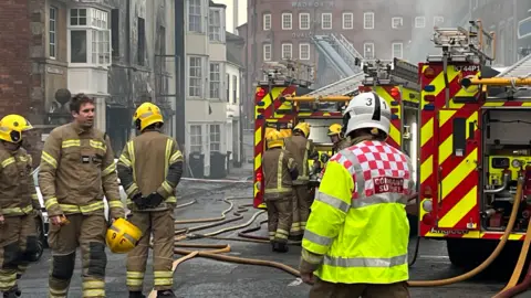 A group of firefighters at the scene of a fire in Wiltshire. They are standing outside the building next to a fire engine. The air is thick with smoke.