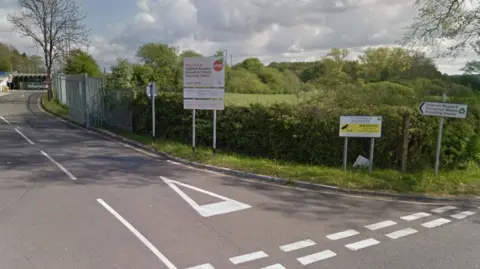 Google The entrance to the Leighton Buzzard Household Waste Recycling Centre with signs on the outside. There is another sign warning it is an offence to fly tip. There is a green field flanked by hedges and trees to the right of the entrance.