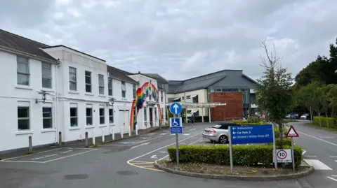 The outside of one of the entrances to Guernsey's hospital. It is a white building with a large rainbow painted on it. There is a silver car parked outside the front of the building. 