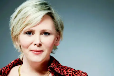 PEGS Michelle has short blonde hair and is wearing a red top with black leopard print on it, and two gold necklaces. 