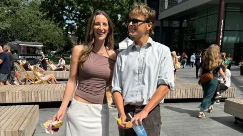 A woman in an off-the-shoulder top, flowy skirt and flat shoes stands next to a man in a shirt, trousers and stomper shoes
