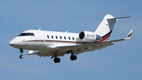 NurPhoto via Getty Images A white Bombardier Challenger 650, with red and blue horizontal stripes, seen against a blue sky