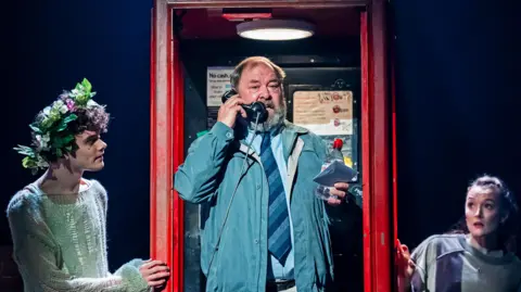Actors on a stage. A middle-age man in a blue jacket, shirt and tie stands in a an old-fashioned red-telephone box, a young man wearing a wreath of flowers and ripped white sweater stands to his right, while a young woman in a jumper stands to his left. 