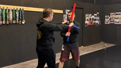 Two men engage in a practice bataireacht fight using red and blue foam sticks. They are stood on black padded mats in the middle of a martial arts studio. Behind them is a wall of different medals and photographs associated with the club.