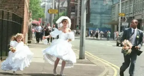 Bride-to-be Mandy Hudson wears a white wedding dress and has a veil on her head. Her bridesmaid, a little girl, is to her right. Both have a panicked look on their faces. A man wearing a grey suit and carrying a bouquet of flowers is to her left. Behind them, several others are running towards the camera.