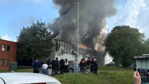 Splash Blinds Aylesbury A crowd of people gather on a grassy bank as they watch a grey building with orange flames on the roof. A large plume of dark smoke is rising into the blue sky.