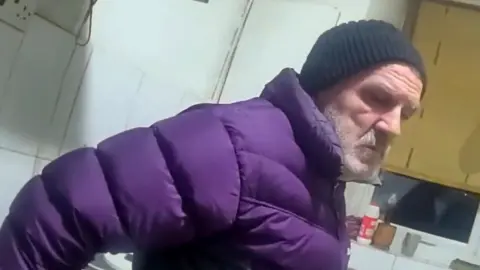 Metropolitan Police Still from police bodycam footage of Eugeniu Neamtu wearing a purple puffer jacket and black beanie hat. He has a grey beard and is standing in a room
