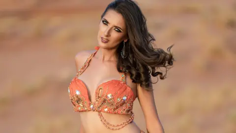 Lizzie Abou Chedid A woman with long dark curled hair, wearing an ornate orange coloured jewelled top, her stomach on show. She is looking directly at the camera.