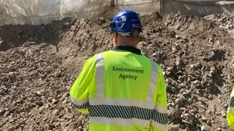 A worker in an Environment Agency fluorescent jacket and a helmet inspects a mound of rubble and soil.