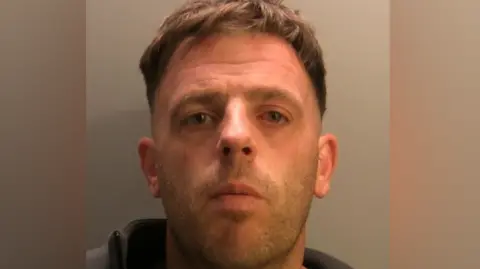 Cumbria Police Daniel Lucas looks straight into the camera in a police mugshot. He has a scar on his right cheek and a cropped short-back-and-sides haircut. He has a small wound on his forehead and is wearing a grey zipped hoodie.