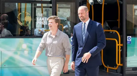 Prince William gets off a tourquise coloured bus wearing a suit and tie