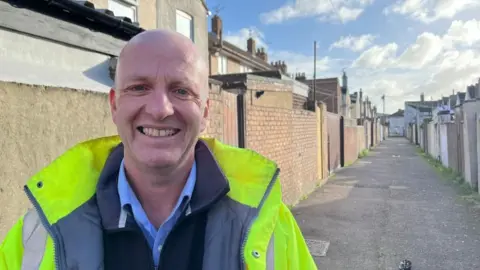 Andrew Turner/BBC James Wilson, wearing a high visibility jacket, black fleece and blue shirt. He smiles and stands in an alley where there are no trash cans and dumps.