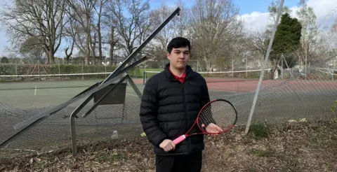 A man with dark hair and wearing a black jacket and trousers, red T-shirt and holding a tennis racket stands in front of the mangled fence with tennis courts behind it, 
