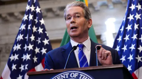 Reuters A man in a suit and a blue tie speaks in front of a podium, with American flags in the background.